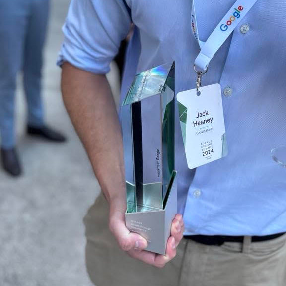 A man in a light blue button-down shirt and khaki pants holds a tall, modern clear acrylic award trophy with green accents. He is wearing a Google branded lanyard with a name tag that reads 'Jack Heaney' and 'Google 2024'.